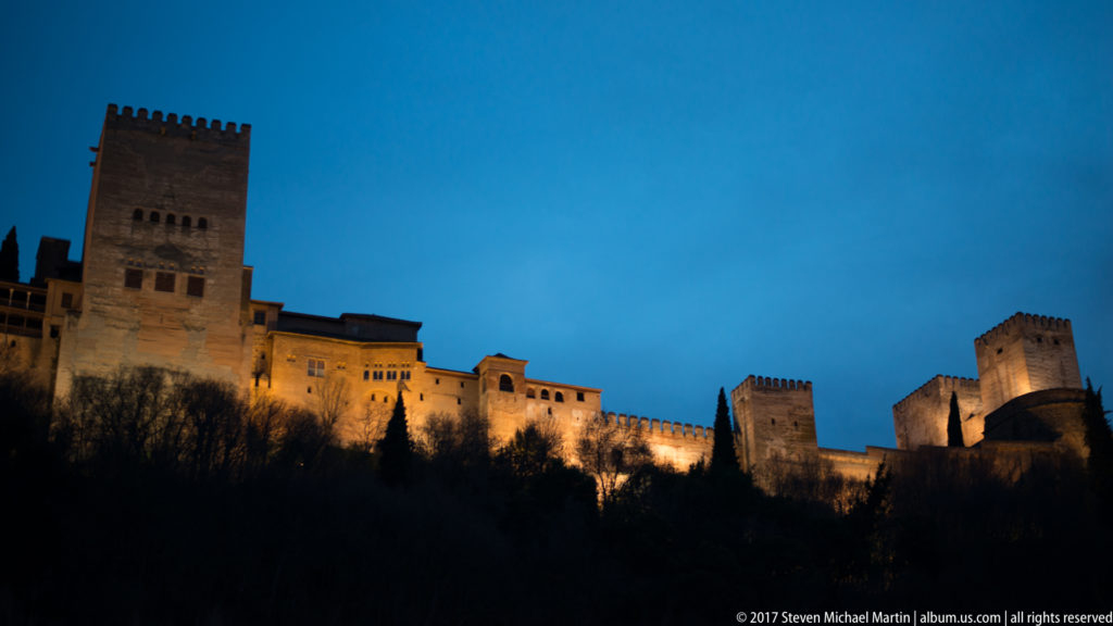 Illuminated Alhambra fortress on a wooded hill at blue hour, with the tall Comares Tower and several lower towers against a deep blue sky.