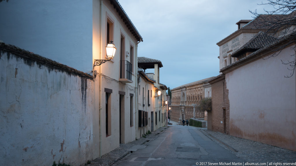 Narrow descending lane inside the Alhambra, bordered by whitewashed walls and traditional lamps, with monumental stone architecture farther ahead.