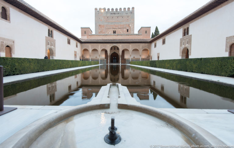 Centered view across the Court of the Myrtles reflecting pool toward the arcaded façade and tall Comares Tower.