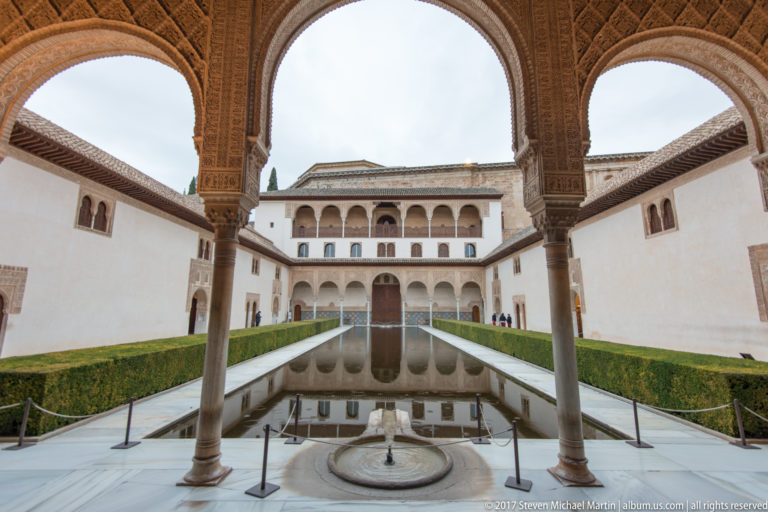 Arcade with slender columns framing the Court of the Myrtles, a small foreground fountain, long pool, hedges, and opposite façade.