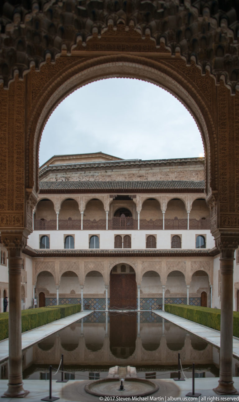View through a decorated arch across the Court of the Myrtles reflecting pool toward a white arcaded façade.