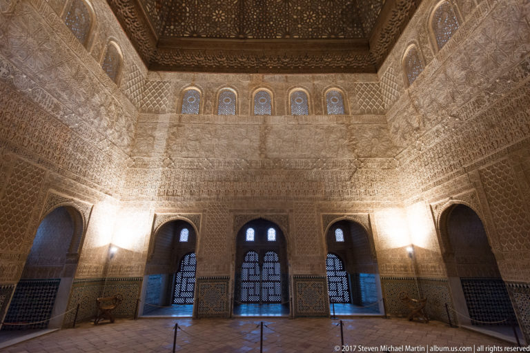 Wide interior of the Hall of the Ambassadors with ornate stucco walls, tall arched windows, and a dark geometric wooden ceiling.