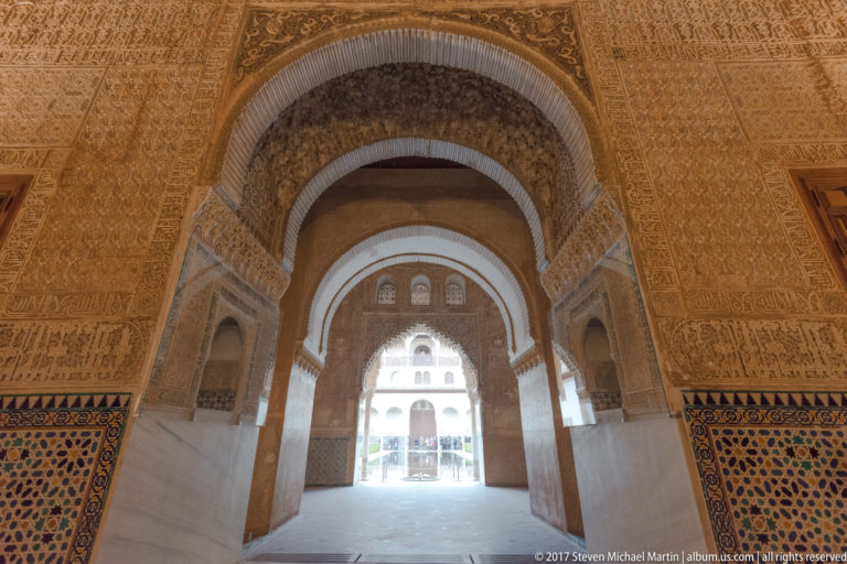 Decorated Nasrid passage with successive arches, geometric tile dados, and a bright courtyard visible at the far end.