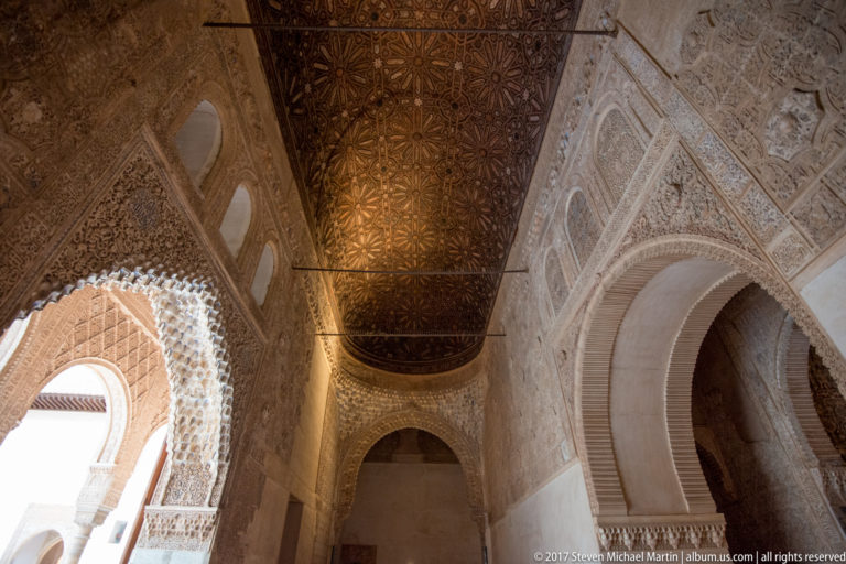 Interior view through ornate horseshoe arches beneath a dark decorated wooden ceiling, with dense carved stucco on the walls.