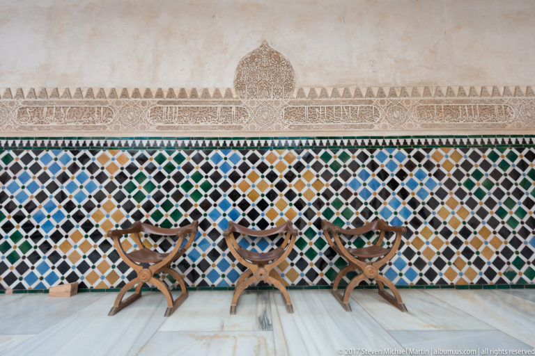 Colorful geometric tile dado beneath pale carved stucco, with three low wooden chairs aligned against the wall.