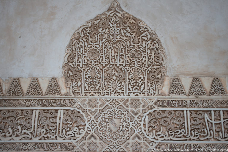 Pointed carved stucco medallion with Arabic calligraphy and dense vegetal ornament above a horizontal decorative frieze.
