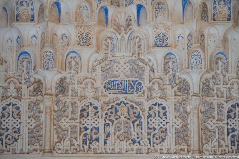 Polychrome Nasrid plaster panel with blue Arabic inscription, muqarnas niches, vegetal carving, and geometric interlace.