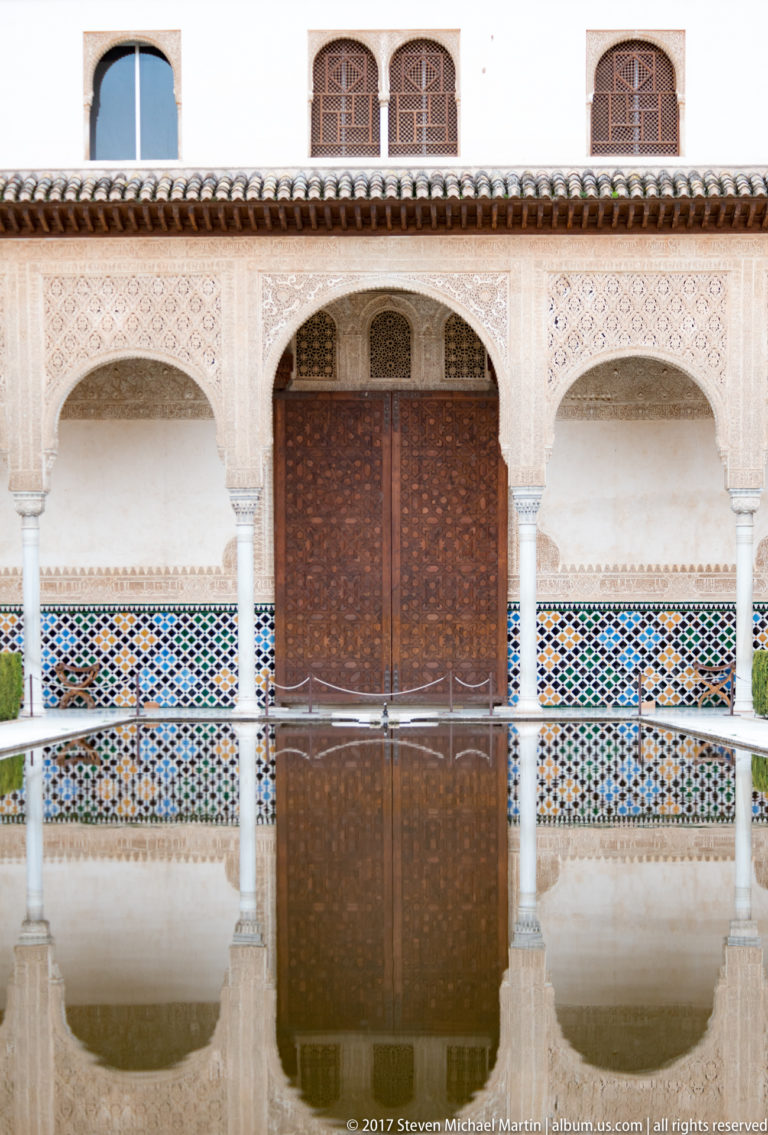 Still reflecting pool before a whitewashed three-arched portico with a central wooden door and colorful geometric tilework.
