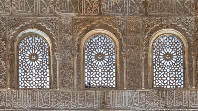 Three narrow arched windows with geometric lattice screens, surrounded by dense carved Arabic inscriptions and stucco ornament.