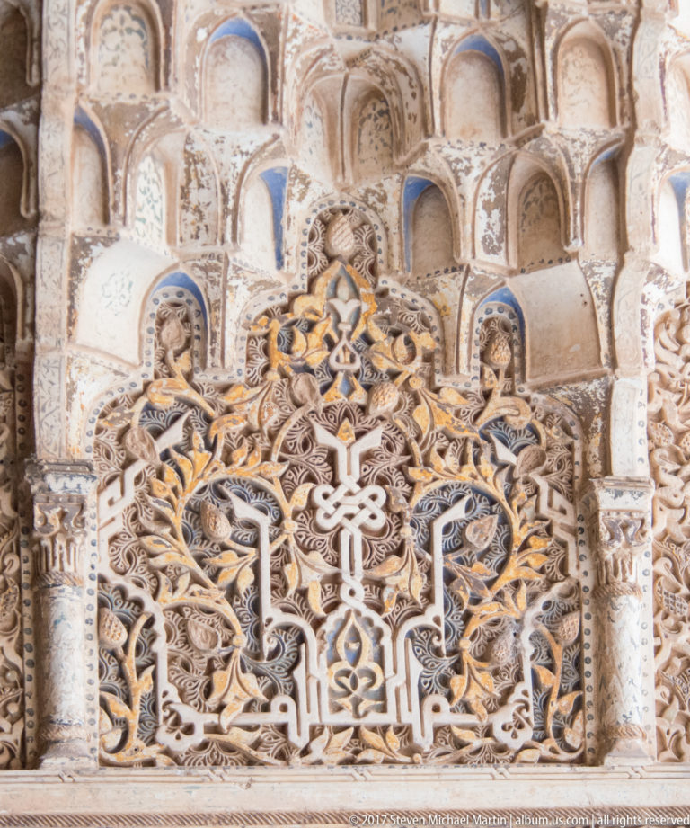 Close view of Arabic calligraphy in carved stucco, framed by geometric ornament and small muqarnas niches above.