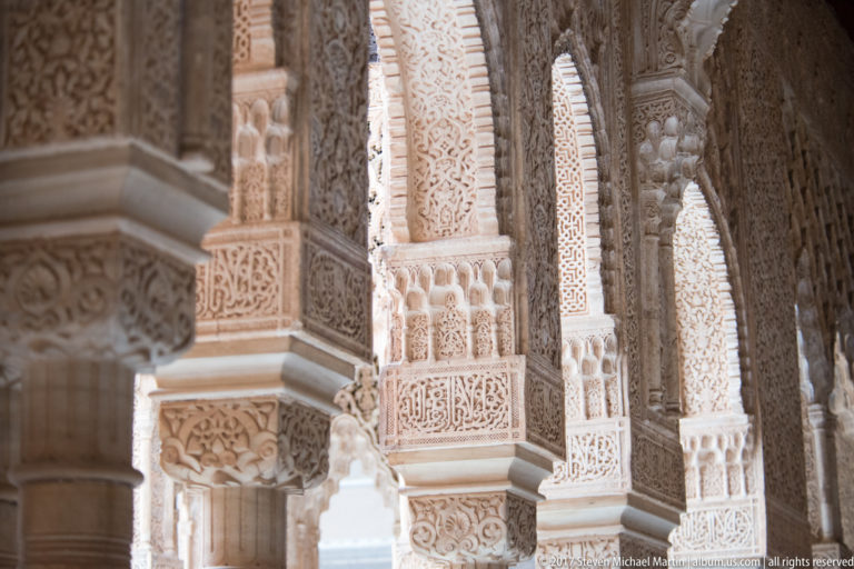 Oblique view through slender columns and carved stucco arches of the Court of the Lions, with bright daylight beyond.