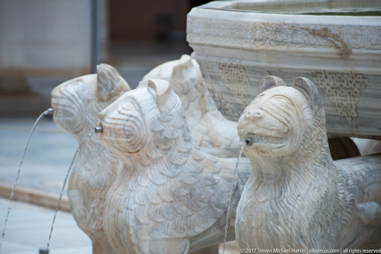 Close view of several carved marble lions beneath the fountain basin, including one lion spouting a narrow stream of water.