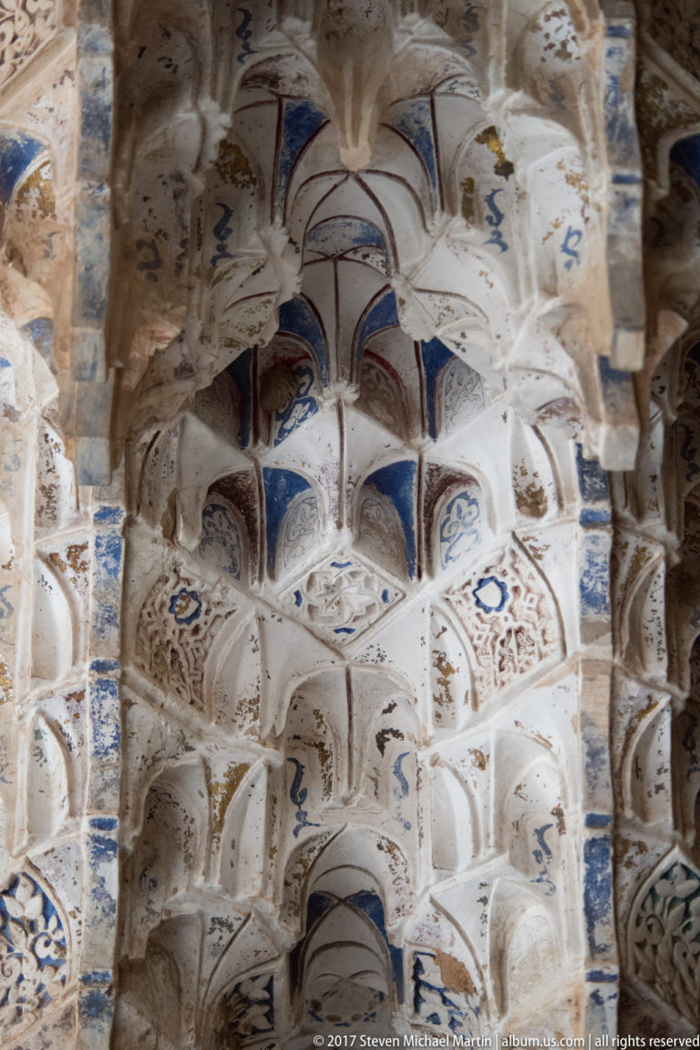 Close view of layered white muqarnas vaulting with blue painted accents and densely carved geometric ornament.