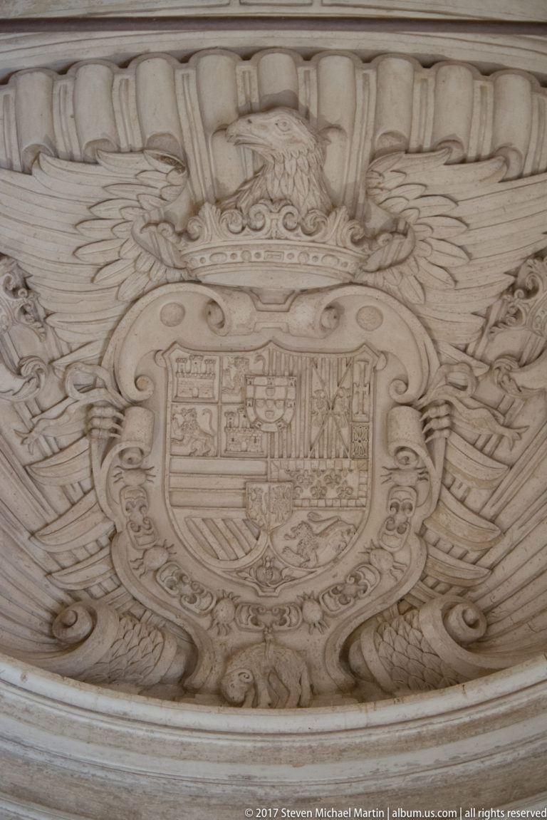 Carved heraldic relief with a crowned shield and elaborate scrolling ornament on pale stone or plaster.