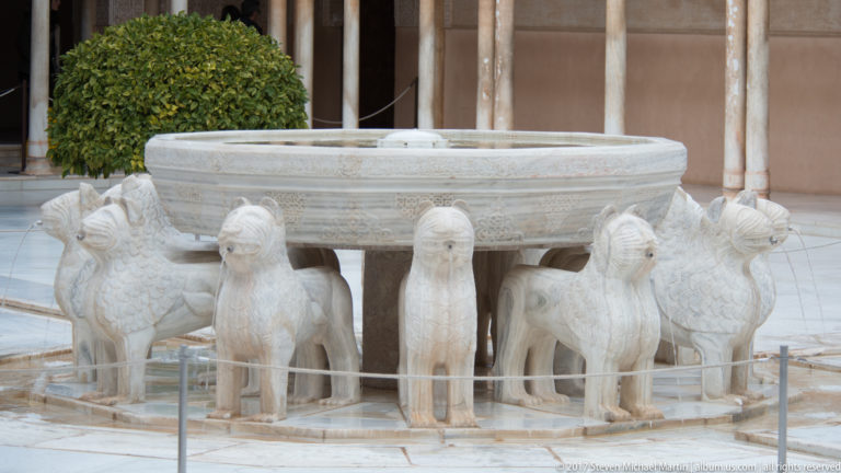 White marble Fountain of the Lions with a carved basin supported by twelve lion figures, several spouting thin streams of water.