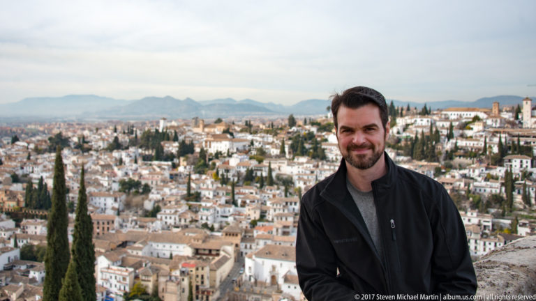 Bearded man in a black jacket posed above Granada, with white Albaicín houses and cypress trees behind him.
