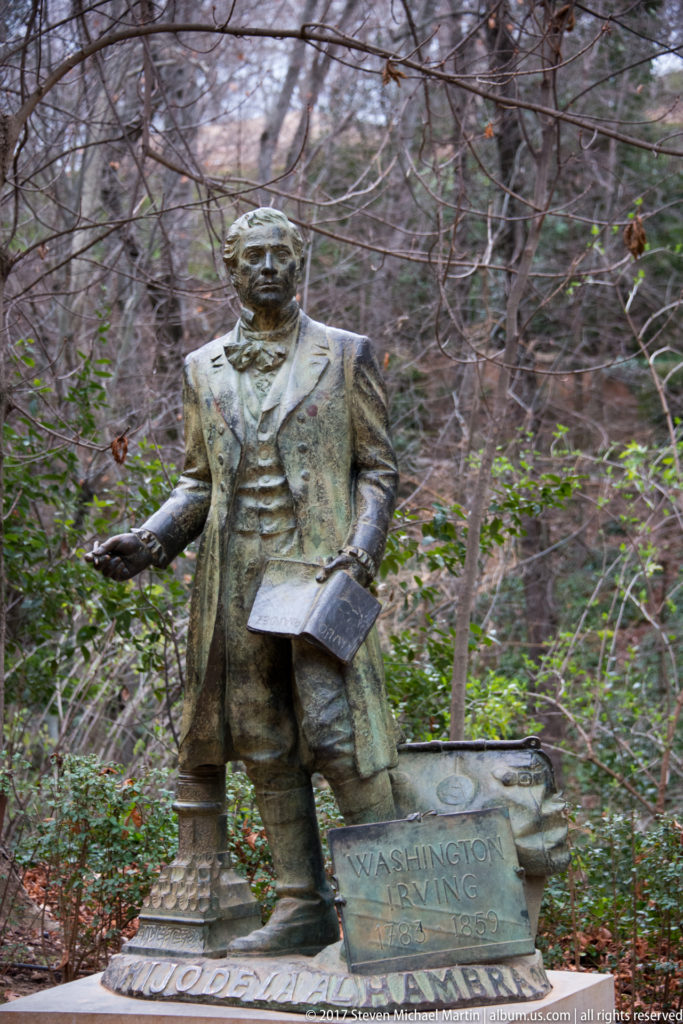 Bronze statue of Washington Irving in nineteenth-century dress standing among trees beside a low inscribed pedestal.