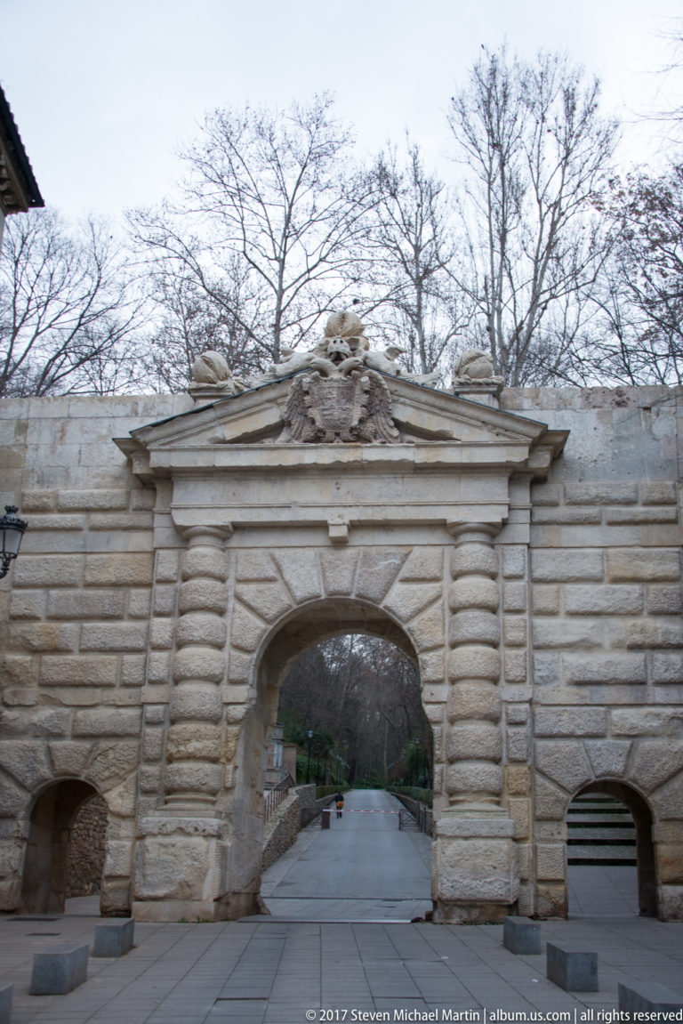 Rusticated stone Puerta de las Granadas with a large central arch, smaller side openings, pediment, and tree-lined road beyond.