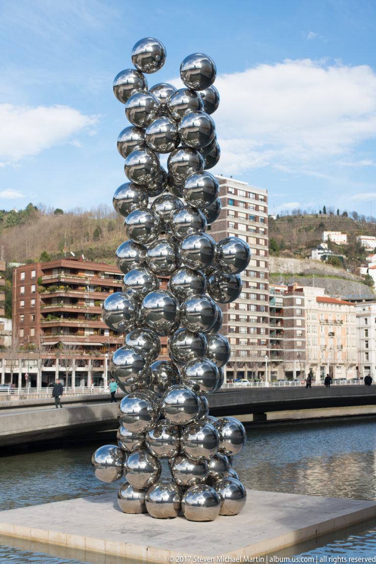 Stacked polished metal spheres form a tall reflective sculpture beside the river, with apartment buildings and hills behind.