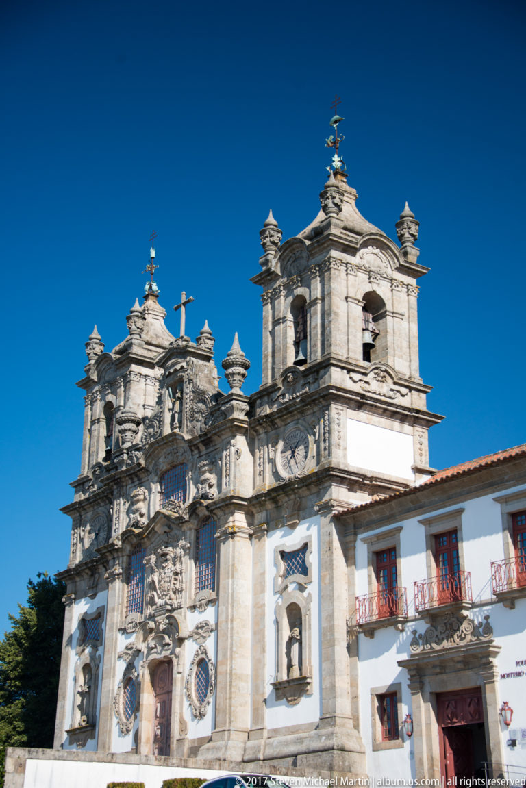 Ornate white church façade with twin bell towers, carved stonework, niches, and a central cross against a deep blue sky.
