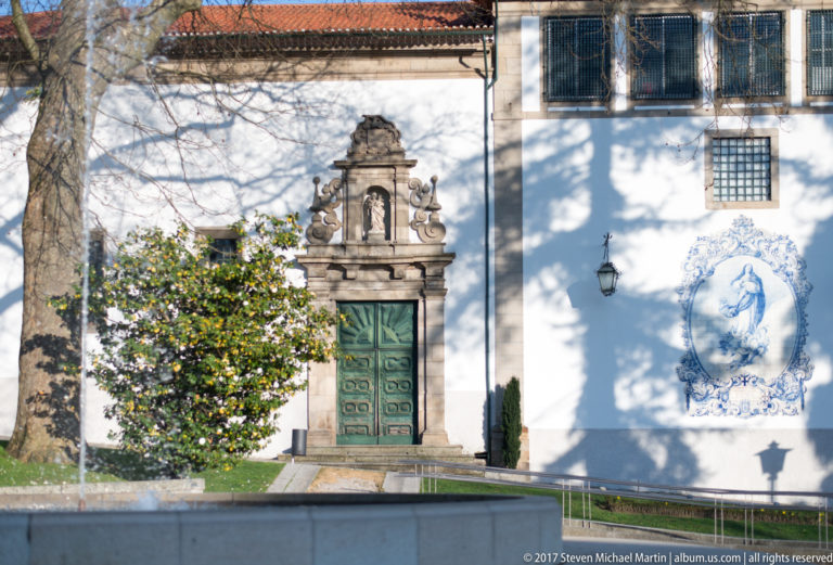 Small chapel with green doors and ornate granite surround beside a large blue-and-white religious azulejo panel.
