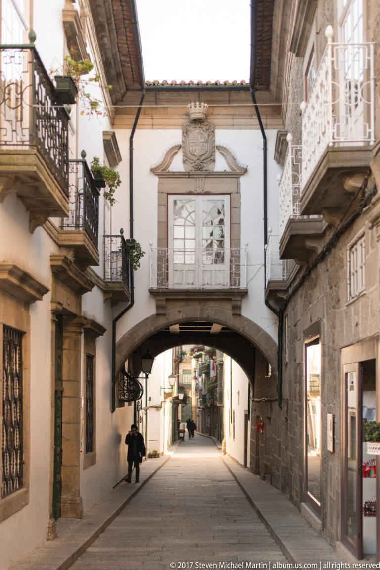 Stone arch spanning a narrow historic street, with balconies, heraldic ornament, white façades, and pedestrians beyond.