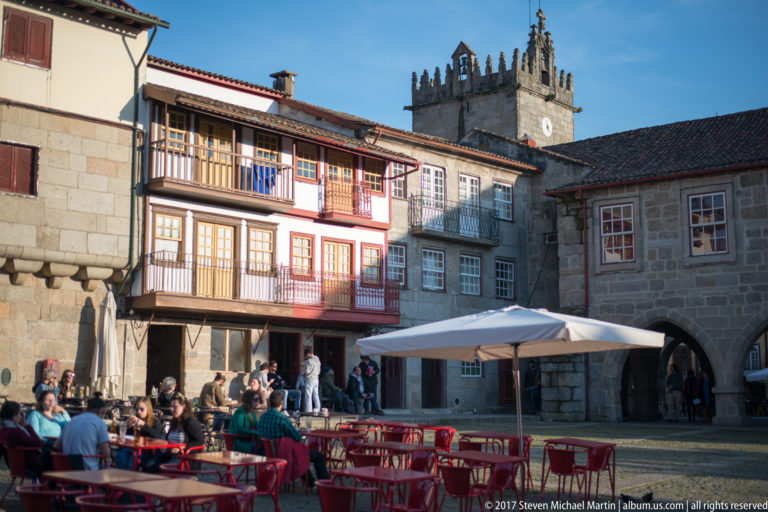 Traditional timber balconies and café seating in Largo da Oliveira with a crenellated church tower rising behind the roofs.