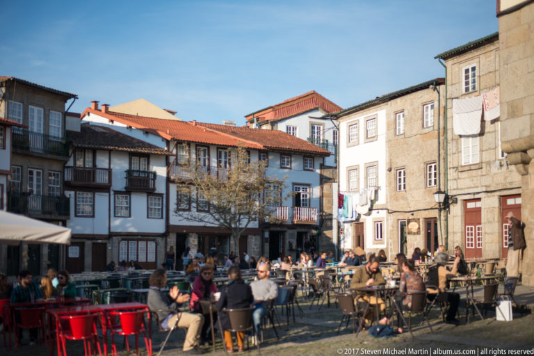 Sunlit historic square with busy café tables, narrow traditional houses, timber balconies, stone façades, and red tiled roofs.