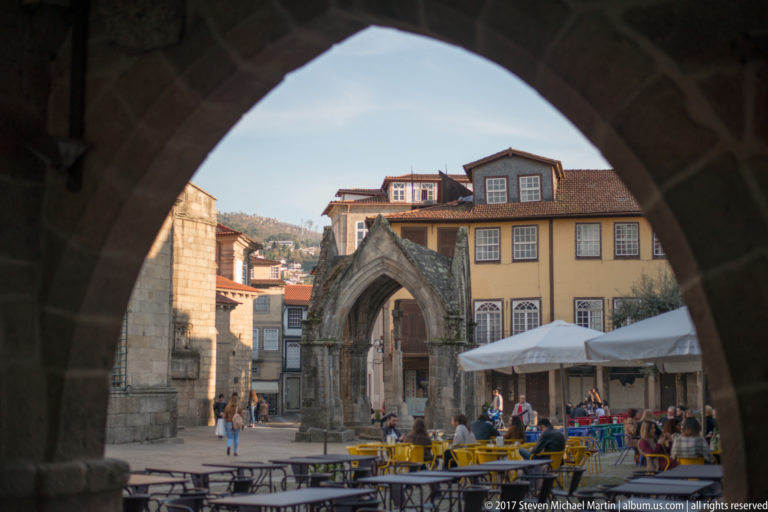 Gothic stone Padrão do Salado with pointed arches framed through a dark archway across a café-filled historic square.