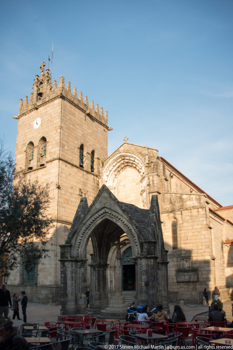 Granite Church of Nossa Senhora da Oliveira with crenellated tower, Gothic portal, large arched window, and café seating in front.