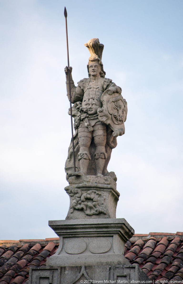 Weathered stone statue of an armored warrior with plumed helmet, spear, and shield standing above a tiled roof.