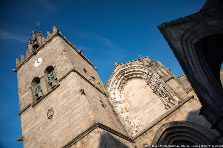 Low-angle view of the crenellated granite tower and carved upper façade of Nossa Senhora da Oliveira against blue sky.