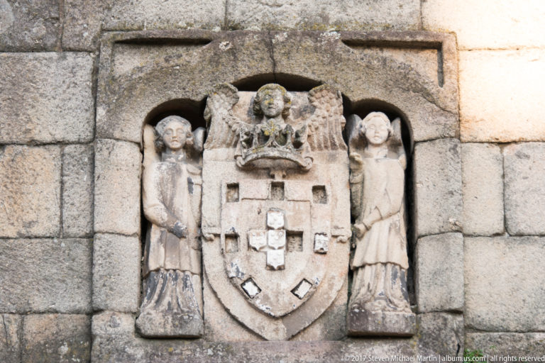 Weathered granite relief of a crowned Portuguese-style coat of arms flanked by two standing carved figures.
