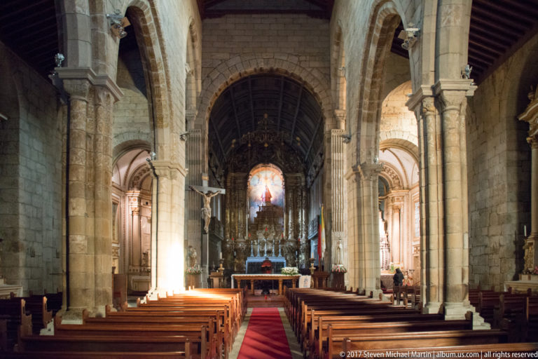 Stone church nave with wooden pews, red runner, pointed arcades, side chapels, and illuminated sanctuary at the far end.