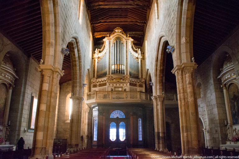 Large ornate pipe organ in a carved pale-and-gilt case above the rear entrance of a granite church nave.