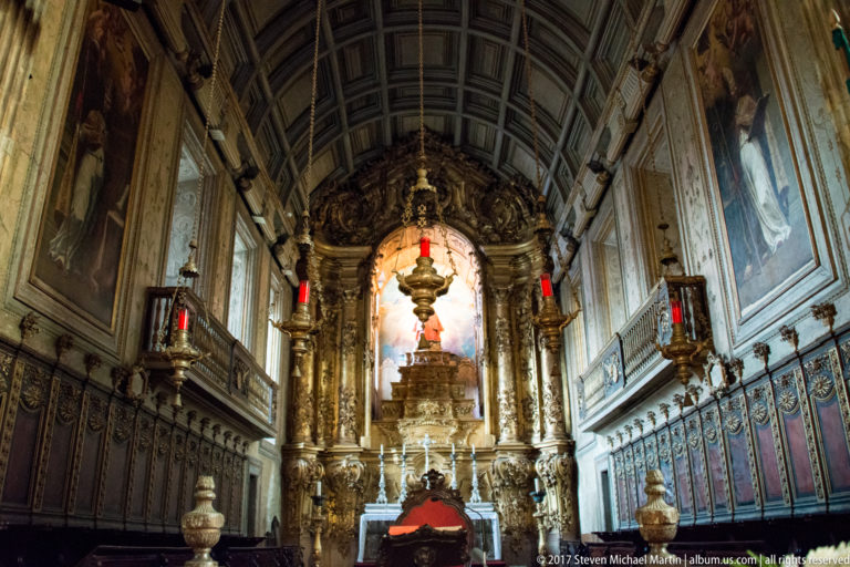 Narrow chapel with richly gilded altarpiece, hanging sanctuary lamp, framed paintings, carved side panels, and painted vaulted ceiling.