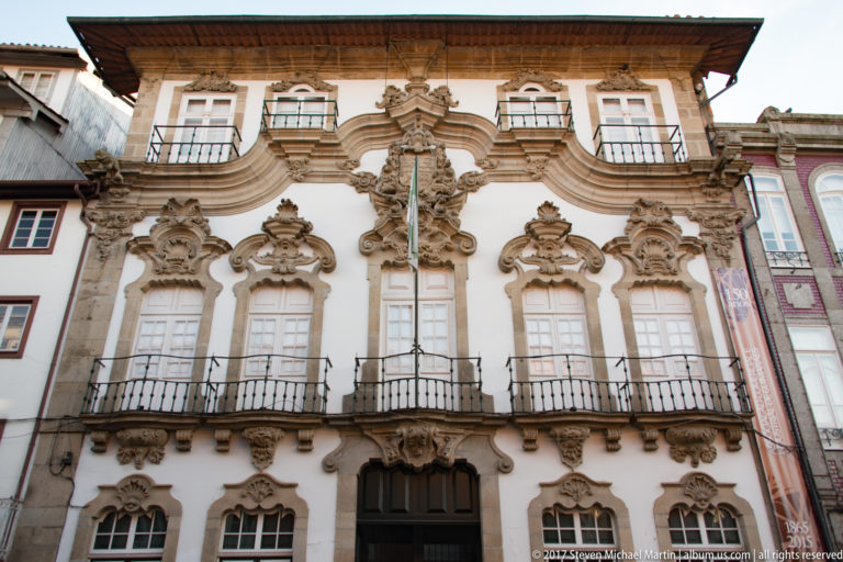 White historic façade with elaborate granite window surrounds, curved pediments, iron balconies, and dense Baroque stone carving.