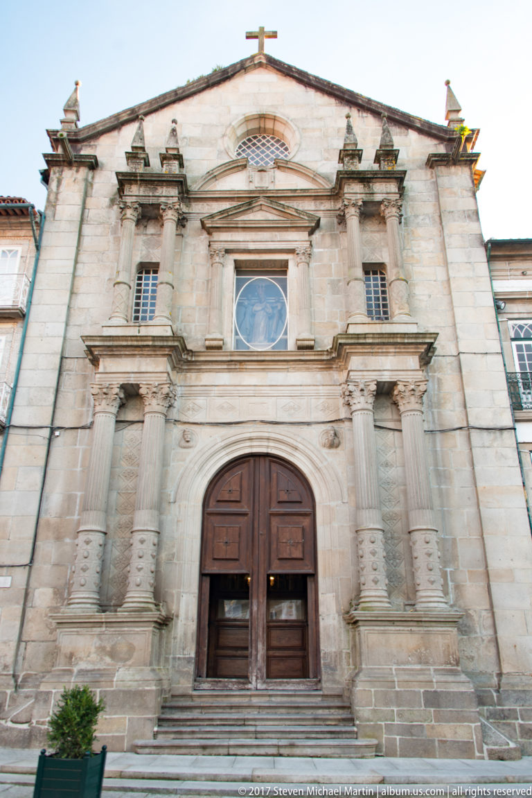 Symmetrical stone church façade with large wooden doorway, circular window, paired pilasters, shallow pediment, and small cross.