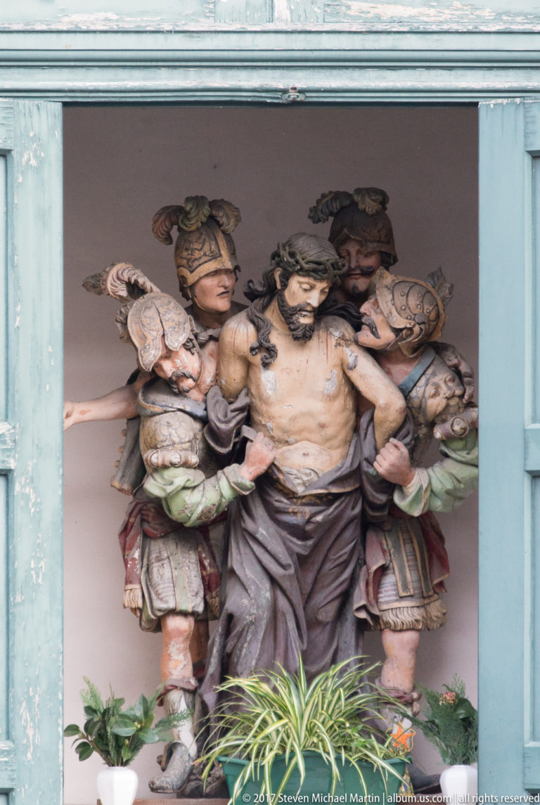 Polychrome sculpture of bound, wounded Christ standing between two armored soldiers in a shallow niche.