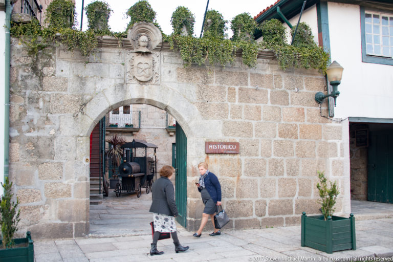 Round-arched granite gateway with greenery above, carved emblem, red “HISTÓRICO” sign, and pedestrians entering a historic passage.