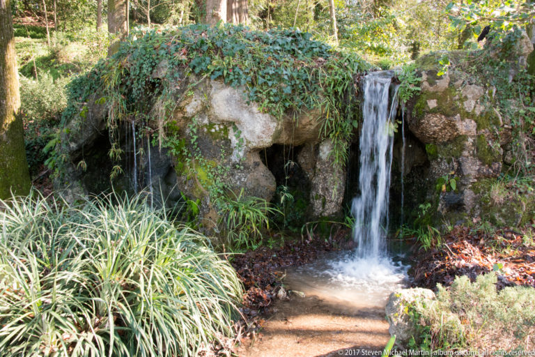 Small waterfall spilling over mossy rocks into a shallow garden basin surrounded by grasses, ivy, and fallen leaves.