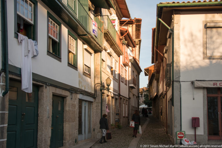 Narrow cobbled Guimarães street lined with stone-and-whitewashed houses, timber balconies, hanging laundry, green doors, and pedestrians.