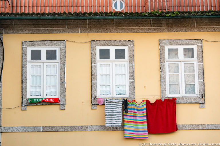 Three granite-framed windows on an ochre wall with colorful laundry, including striped garments and a small Portugal banner, hanging below.