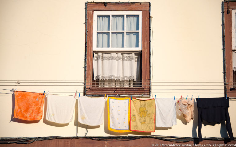 Traditional window with lace curtain above a clothesline holding towels, shirts, underwear, and dark garments against a pale wall.