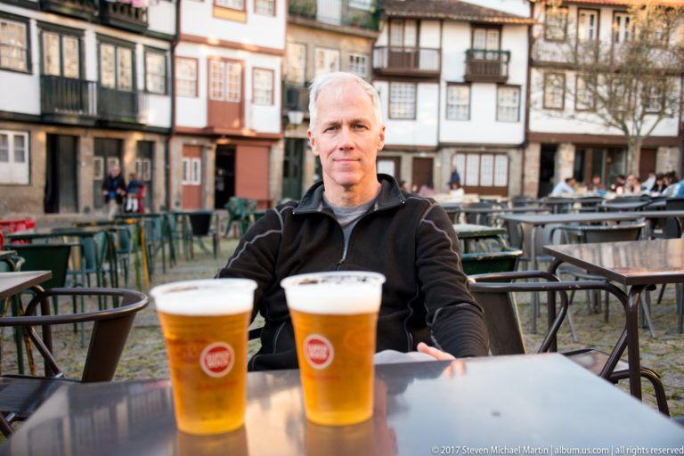 Gray-haired man seated at an outdoor café table with two beers, surrounded by empty chairs and historic façades.