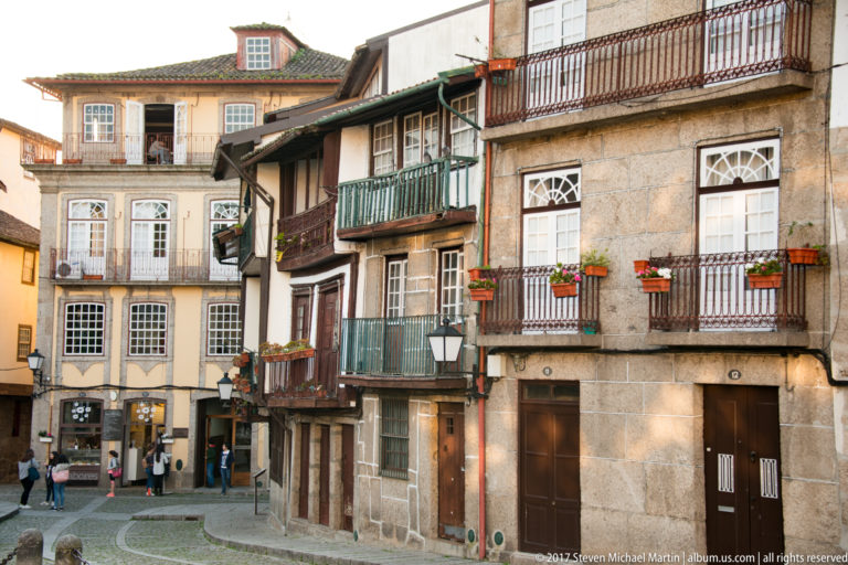 Traditional multi-story houses with granite façades, projecting balconies, flower boxes, green woodwork, and narrow doors around a paved junction.