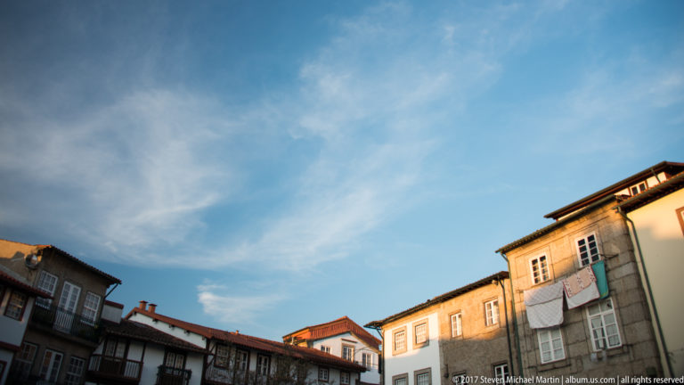 Historic Guimarães rooftops and upper façades catching warm evening light beneath a large blue sky with wispy clouds.