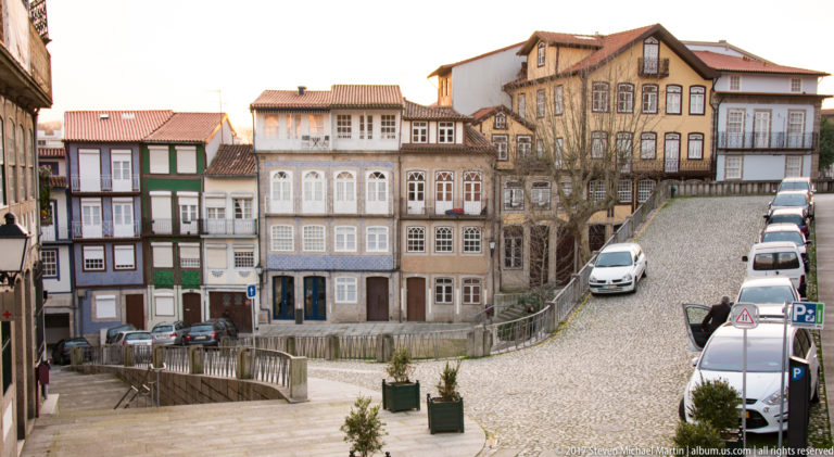Historic houses with glazed balconies and timber trim beside a steep cobbled street lined with parked cars in Guimarães.