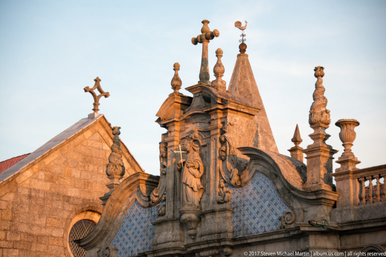 Close view of ornate stone crest, scrolls, urn finials, and pinnacles above blue-and-white tiles beside a Gothic gable.