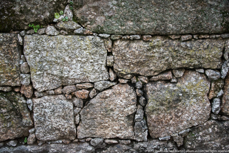 Close-up of weathered granite masonry built from large rough blocks, smaller packing stones, mortar joints, and patches of lichen.